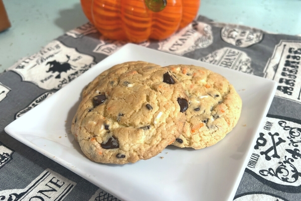 Cookies with orange, black, and white sprinkles and chocolate chips on a white plate with black and white Halloween towel under the plate and part of a glass pumpkin in background